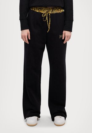 Persona con pantalones de chándal negros holgados con cinturilla decorada en dorado y cordón, camiseta negra y zapatillas blancas, de pie mirando al frente.