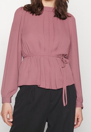 Kvinde iført en mauve langærmet bluse med bindebånd foran og plisseringer, kombineret med løstsiddende sorte bukser.