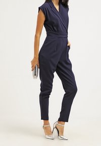 Mörkblå jumpsuit med omslag, korta ärmar och bälte i midjan. Har avsmalnande ben och en slät yta, i kombination med silverfärgade klackar.