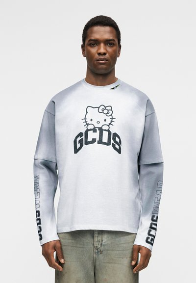 Mies pukeutunut valko-harmaaseen pitkähihainen paita, jossa Hello Kitty -grafiikka ja "GCDS"-teksti, yhdistetty vaaleanharmaisiin farkkuihin.