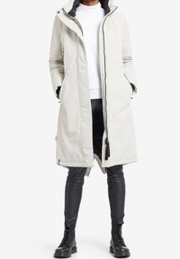 Pitkä, vaaleanharmaa vetoketjullinen takki hupulla, jossa on musta vuori, sivutaskut ja lievästi kapeneva muotoilu. Yhdistetty mustiin leggingsiin ja saappaisiin.