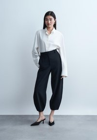 Moteris stovi priešais paprastą sieną, dėvinti baltą pernelyg didelę marškinių palaidinę, juodus aukštos liemens plačiakampius trumpintus kelnes ir juodus smailius aukštakulnius.