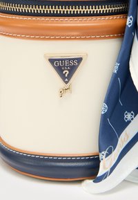 Στρογγυλή τσάντα χειρός σε κρεμ χρώμα με ναυτικό και ταμπά λεπτομέρειες, χρυσά μεταλλικά στοιχεία. Διαθέτει τριγωνικό λογότυπο Guess και ένα διακοσμητικό μπλε φουλάρι.