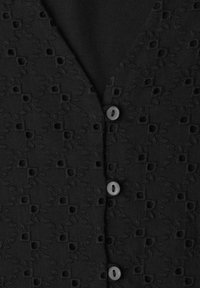 Tissu noir avec motif carré et floral découpé brodé, avec un col en V et trois petits boutons noirs au centre.