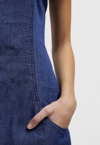 Main placée dans la poche latérale d'une robe en jean bleu sur un fond clair uni.