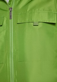 Jachetă verde cu fermoar, cu un finisaj texturat. Prezintă două buzunare la piept cu capac și un fermoar neted în centru.