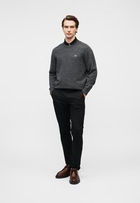 Suéter gris de punto con cuello negro y logotipo en el pecho. Combinado con pantalones negros y zapatos de cuero marrón. Hombre posando frente a un fondo liso.