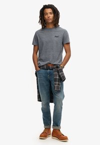 Grijze katoenen t-shirt met een ronde hals, blauwe spijkerbroek en bruine leren laarzen. Een geruite flanellen shirt is om de taille gebonden.