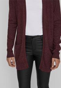 Burgundy neulekarhu, jossa on auki oleva etuosa, joustinneuleiset hihansuut ja kaksi etutaskua, yhdistettynä mustiin slim-fit-housuihin.