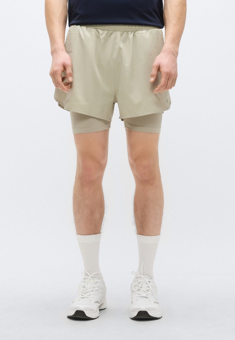 Pantaloni scurți atletici bej peste pantaloni scurți cu compresie ușoară, cu talie elastică, textură netedă și două buzunare laterale. Asortați cu adidași albi.