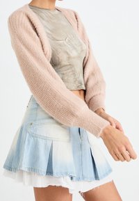 Donna che sistema la manica indossando un cardigan beige in peluche, un top cropped verde oliva e una gonna di jeans pieghettata azzurro chiaro con orlo sfilacciato bianco.