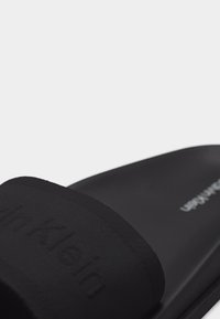 Mule noire Calvin Klein avec large bride en tissu et semelle intérieure rembourrée, présentée sur un fond uni.