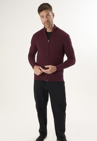 Bordeauxfarbenes, gestricktes Cardigan mit Reißverschluss, gerippten Kanten und langen Ärmeln, kombiniert mit schwarzen Hosen und Schuhen vor einem neutralen Hintergrund.