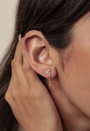 Goldene Ohrhänger mit funkelnden klaren Edelsteinen, die ein florales Design und Kettendetails aufweisen, nahe am Ohr einer Person positioniert.