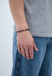 Karoliukas su juodais akmenimis ir auksinėmis detalėmis, su maža kryžiaus detalė. Dėvimas ant rankos, prieš pilką marškinėlį ir džinsus.