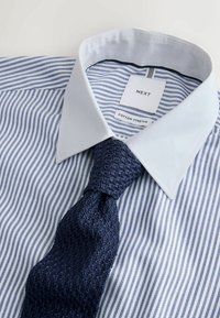 Chemise à rayures bleu et blanc avec un col blanc et une cravate en tricot bleu marine texturée, étiquette de marque visible à l'intérieur du col.