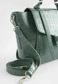 Sac à main en cuir vert à motif crocodile, avec une longue bandoulière détachable et des fermoirs en métal argenté sur fond blanc.