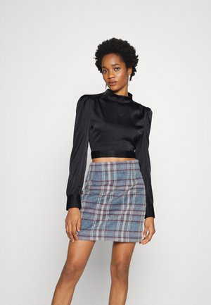 Top cropped em cetim preto com mangas bufantes longas e gola alta, combinado com uma saia justa em padrão xadrez nas tonalidades de azul, cinza e vermelho.
