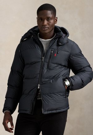 Homme portant une doudoune noire avec capuche, un pull gris clair, un pantalon noir et une montre au poignet gauche, debout devant un fond uni.