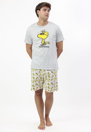 Hombre joven de pie descalzo, vistiendo una camiseta gris con un personaje amarillo de Woodstock y pantalones cortos blancos a juego con el patrón de Woodstock.