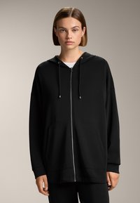 Felpa nera con zip realizzata in materiale morbido, dotata di cappuccio, due tasche frontali e dettagli con cordino. Vestibilità comoda con polsini a coste.