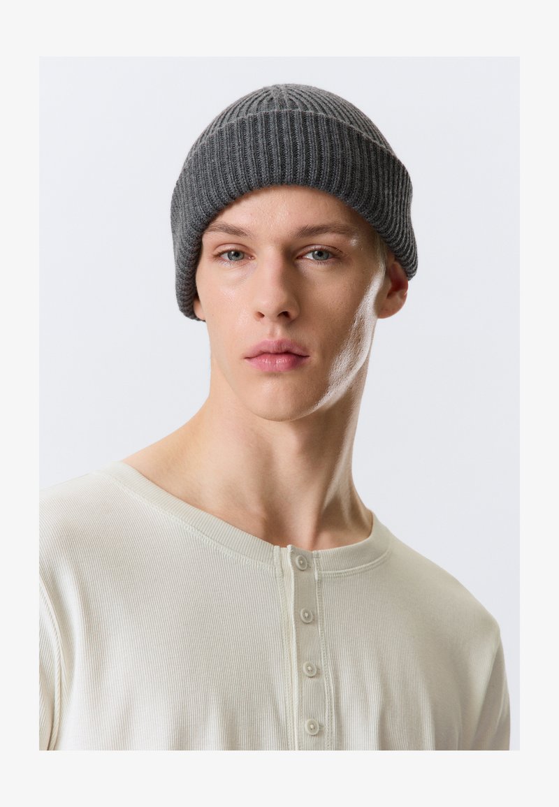 Grijze geribbelde gebreide muts met een strakke pasvorm, gedragen door een persoon in een lichtbeige shirt met lange mouwen en een knoopsluiting aan de hals.