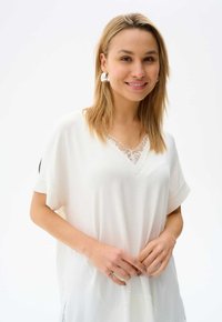 Witte tuniektop met een v-hals en kantdetails, korte mouwen en een lichtgewicht textuur. Model staat met de handen gekruist bij de taille.