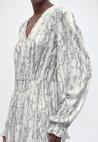 Femeie purtând o rochie cu mâneci lungi, decolteu în V, manșete adunate și model de șarpe gri-alb pe un material ușor.