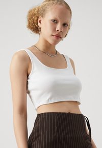 Hvit stripete crop-top med firkantet U-hals, kombinert med mørkebrune høyt midje bukser med pinstriper. Glatt tekstur, tettsittende design.