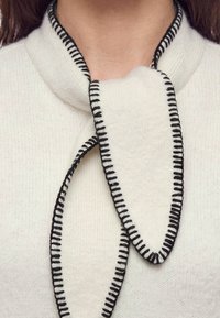 Gros plan d'une écharpe blanche tricotée avec des bords de points noirs enroulée autour du cou d'une personne par-dessus un pull blanc.