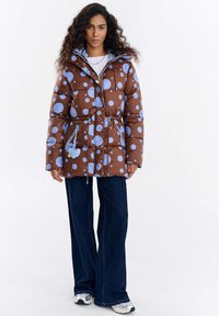 Brązowa kurtka puchowa z dużymi niebieskimi groszkami, regulowanym ściągaczem w talii i dwoma przednimi kieszeniami na zamek. Połączona z szerokimi niebieskimi dżinsami.