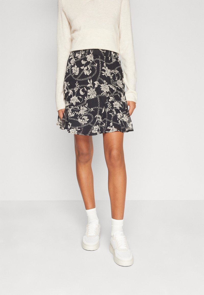 Femme portant une mini-jupe noire à fleurs, un pull blanc à manches longues, des chaussettes blanches et des baskets blanches, debout devant un fond uni.