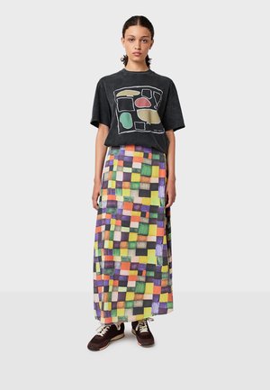 Una joven se encuentra de pie vistiendo una camiseta gráfica negra, una larga falda a cuadros multicolor y zapatillas oscuras con cordones y suelas blancas.