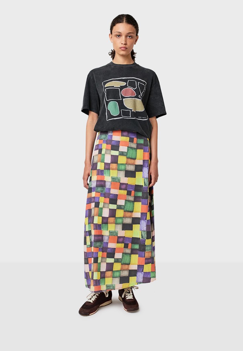 Une jeune femme se tient debout, portant un t-shirt graphique noir, une longue jupe à carreaux multicolores et des baskets sombres avec des lacets et des semelles blanches.