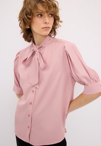 Blusa rosa pallido realizzata in tessuto morbido, con colletto a fiocco, maniche a sbuffo e chiusura con bottoni. Texture liscia con vestibilità rilassata.