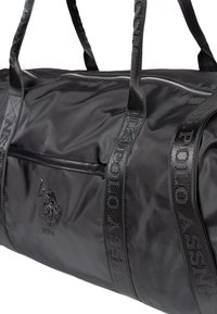 Černá sportovní taška vyrobená z tkaniny, s dvěma popruhy, zipovým uzavřením a logem na přední straně s texturovanými popruhy.