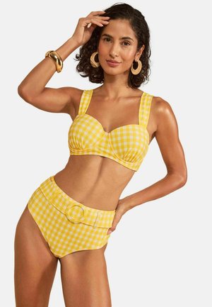 Frau mit kurzen lockigen Haaren, die einen gelb-weiß karierten Bikini mit hoher Taille, große Creolen und ein goldenes Armband trägt, posiert mit der Hand am Kopf.