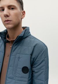 Chaqueta acolchada azul con un cuello alto, que presenta un parche circular negro en el pecho y un bolsillo con cremallera en el lado izquierdo.