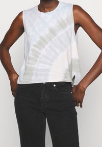 Bavlněný bezrukávový crop top s tie-dye vzorem v světle modré a béžové barvě, se bočním rozparkem a kulatým výstřihem. V kombinaci s černými džíny.