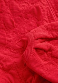 Tissu matelassé rouge avec des motifs de cœur cousus et un bord-côtes, probablement une partie d'une manche de pull ou de sweat-shirt.