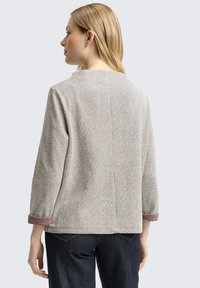 Vrouw met blond haar die een lichtgrijze geribbelde trui met opgerolde mouwen en donkerblauwe spijkerbroek draagt, van zich afgekeerd tegen een effen achtergrond.