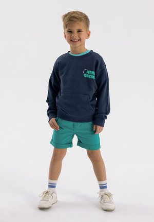 Námornícky mikina s teal logom, spárovaná s teal šortkami. Biely tenisky s modrými a sivými pruhmi na ponožkách. Pohodlný strih a dizajn.