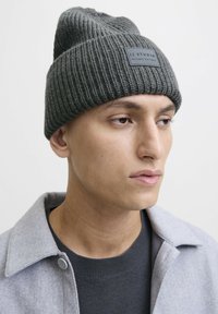 Jonge man met een grijze gebreide beanie met een "JJ Studio" patch, een grijze jas en een donkere trui, kijkt naar rechts tegen een effen achtergrond.