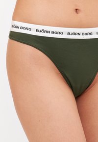 Olive green bikini brief with a white elastic waistband featuring black "BJÖRN BORG" branding. Soft fabric and minimal coverage design.