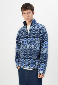 Blå og grå fleece pullover med halv-lynlås krave, prydet med geometriske mønstre af diamanter og zigzag. Blød tekstur, afslappet pasform.