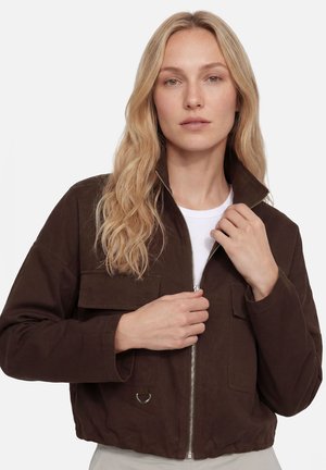 Femme blonde portant une veste courte marron foncé sur une chemise blanche, en train de fermer la fermeture éclair à l’avant, sur un fond uni.