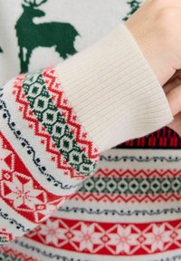 Maglione lavorato a maglia con una varietà di motivi in rosso, verde e blu, con polsini a coste color crema. Include renne e fiocchi di neve.