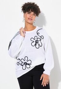 Vit stickad tröja med svarta abstrakta blommönster i applikationer, långa ärmar, ribbade manschetter och en lös passform, i kombination med svarta byxor och en silverhandväska.