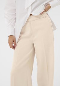 Pantalon beige à jambes larges en tissu doux, doté d'une taille plate et de poches latérales, associé à une chemise blanche à manches volumineuses.