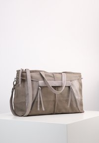 Sac fourre-tout en cuir et en daim marron clair avec deux poignées, une bandoulière amovible et des poches zippées à l'avant sur une surface blanche.